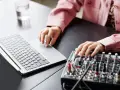 Osoba z tatuażami na dłoniach reguluje suwaki na mikserze, obok laptopa. Dowiedz się, jak podłączyć mikser do laptopa.