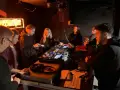 Grupa młodych ludzi przy konsoli DJ-skiej, uczących się, jak zostać DJ-em. Neonowy napis w tle.
