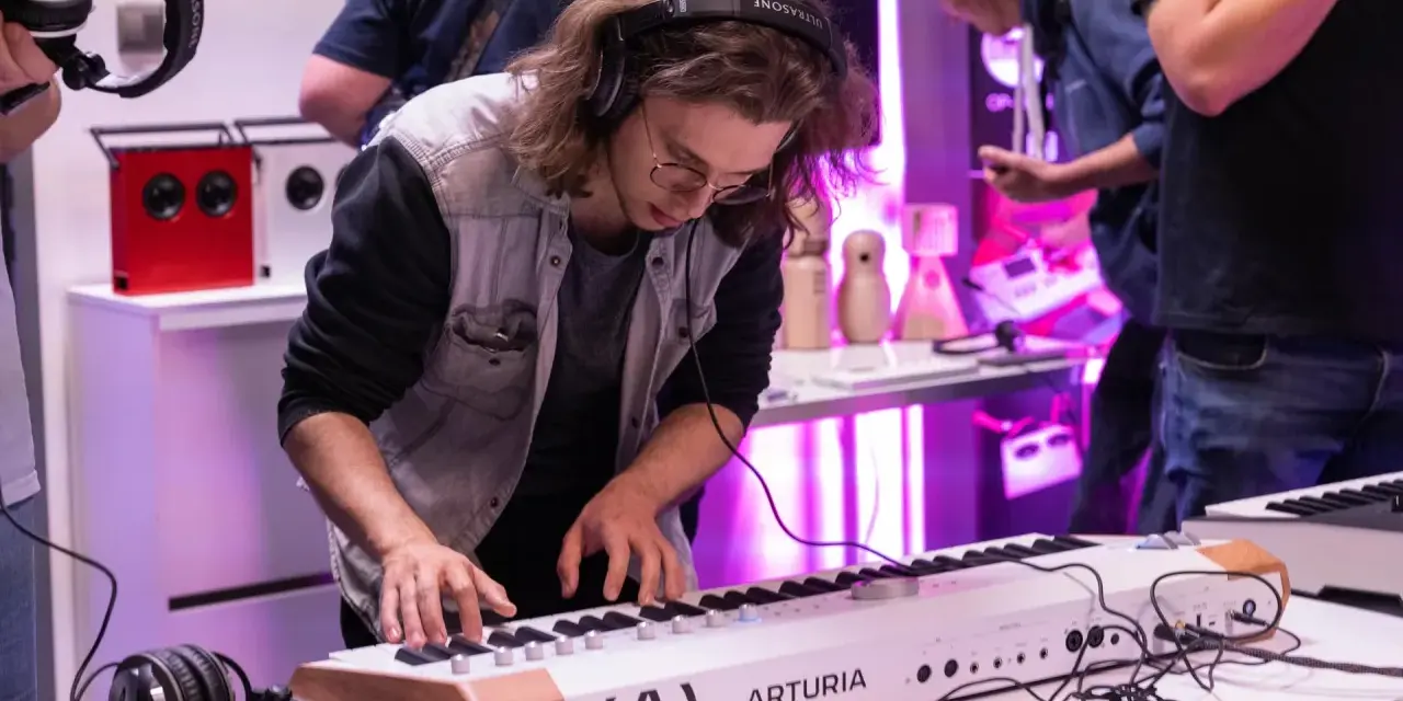 Młody mężczyzna w okularach i słuchawkach ćwiczy na keyboardzie Arturia. To część kursu produkcji muzycznej, gdzie można poznać tajniki tworzenia dźwięków.