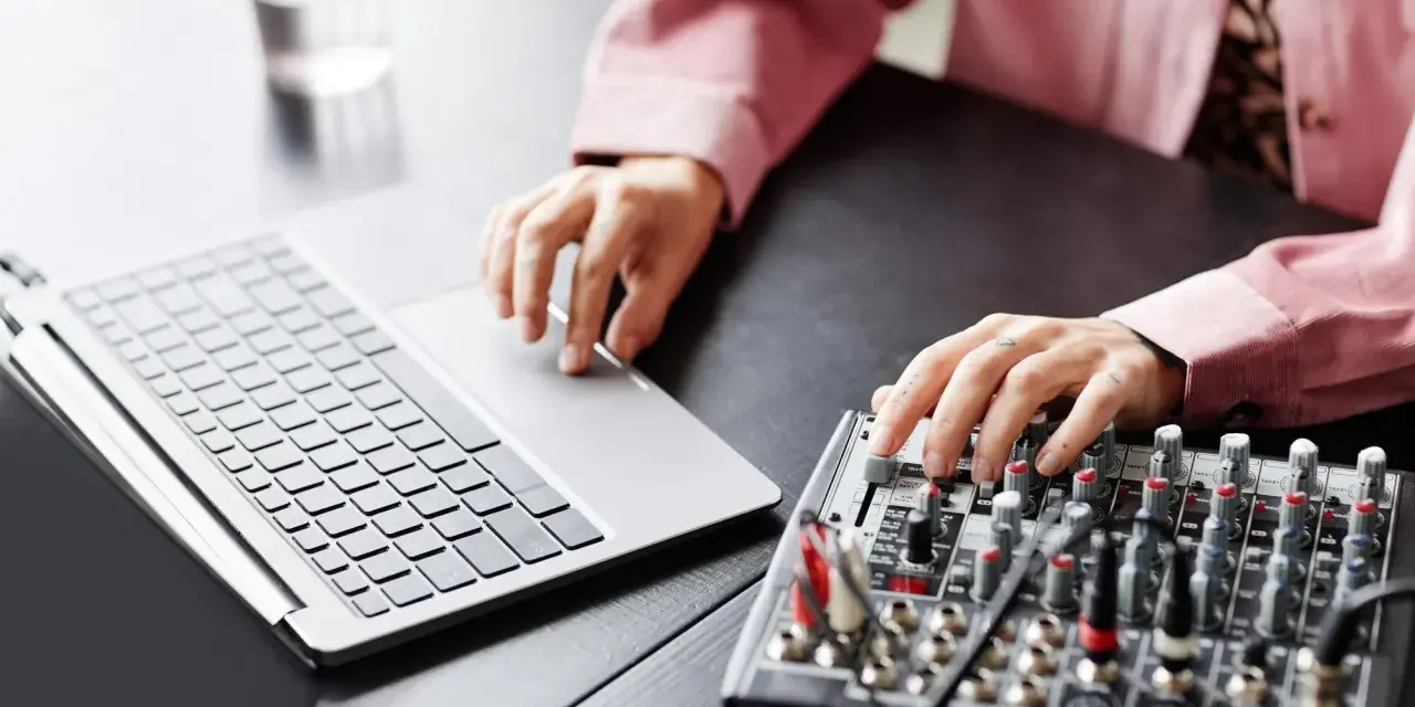 Osoba z tatuażami na dłoniach reguluje suwaki na mikserze, obok laptopa. Dowiedz się, jak podłączyć mikser do laptopa.