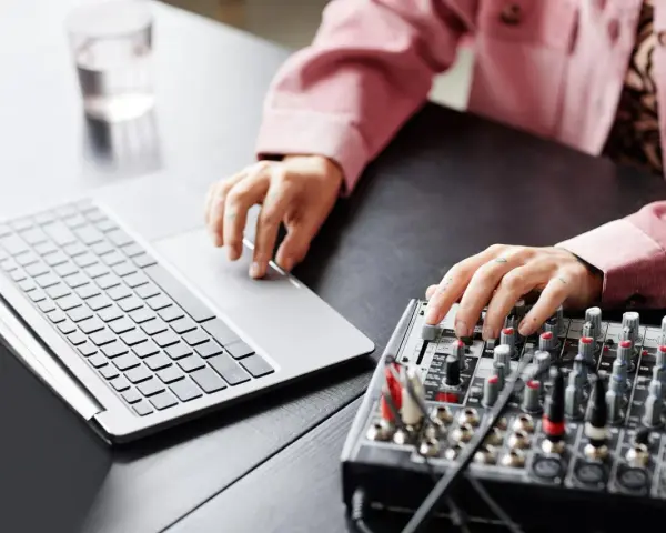 Osoba z tatuażami na dłoniach reguluje suwaki na mikserze, obok laptopa. Dowiedz się, jak podłączyć mikser do laptopa.