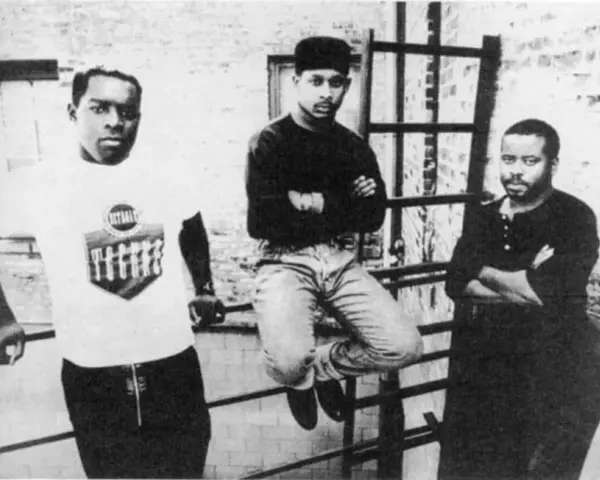 Trzech mężczyzn, pionierzy detroit techno, pozuje na tle ceglanej ściany.