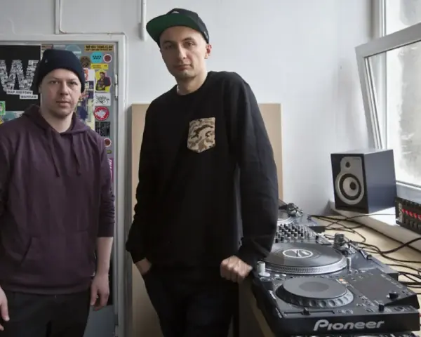 Dwóch mężczyzn przy sprzęcie DJ-skim. Jeden w bordowej bluzie z kapturem, drugi w czarnej bluzie z naszywką. To ich sposób na pokazanie, co to stereo.