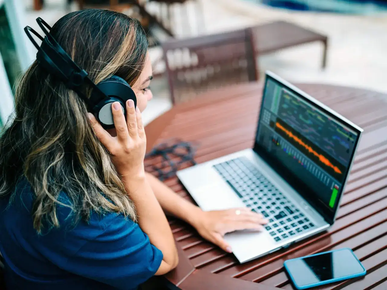 Kobieta w słuchawkach uczy się produkcji muzyki na laptopie, siedząc przy stoliku na zewnątrz.