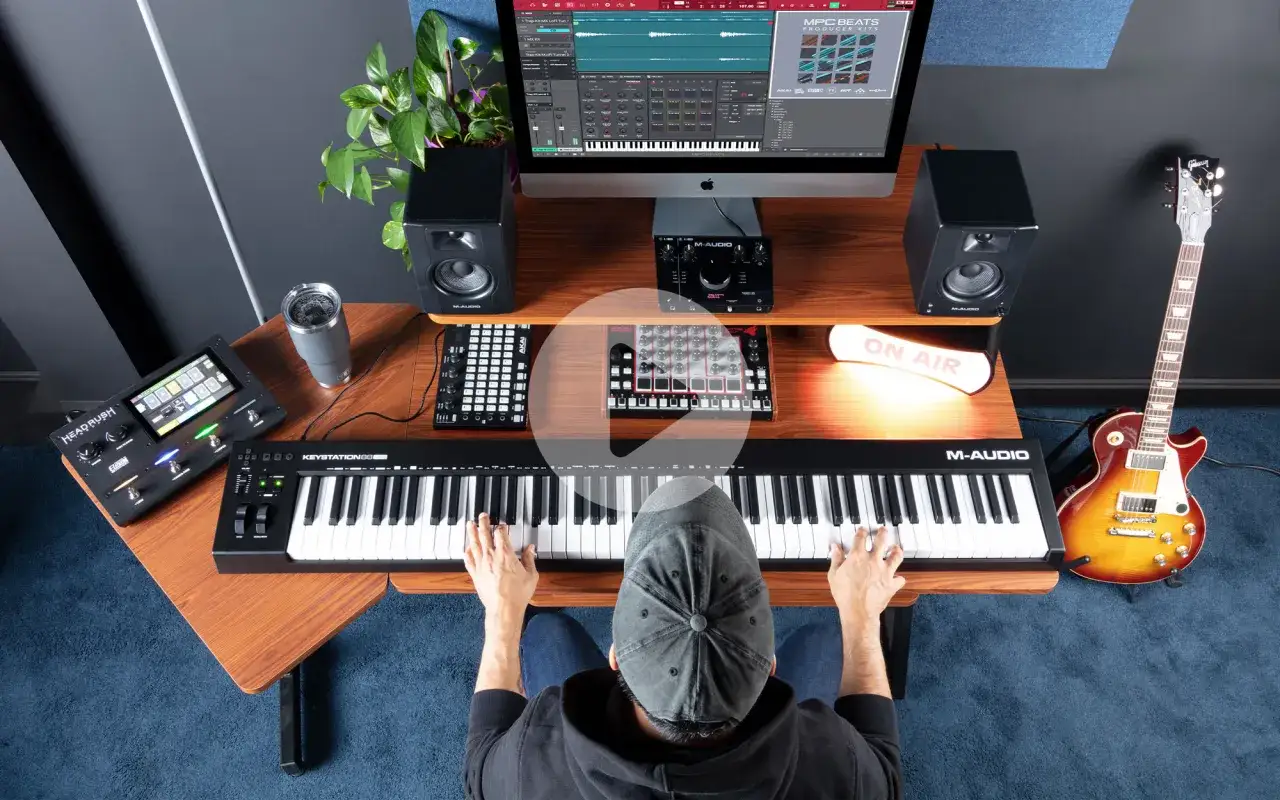 Osoba gra na klawiaturze M-Audio Keystation 88 MK3 w domowym studiu nagraniowym, obok gitary i sprzętu do produkcji muzyki.