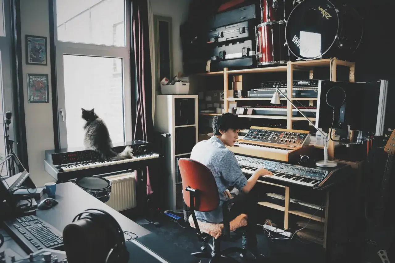 Muzyk tworzy dźwięki na syntezatorze Yamaha DX7, podczas gdy kot obserwuje go z okna.